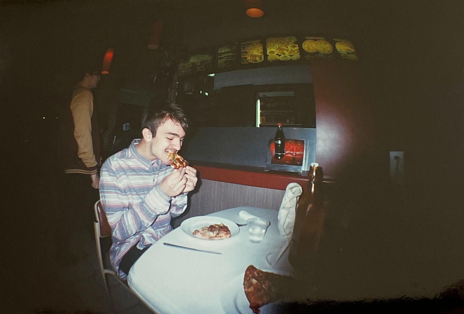 A fish-eye style photo of the author as a teenager, sitting at a table and biting into a slice of pizza