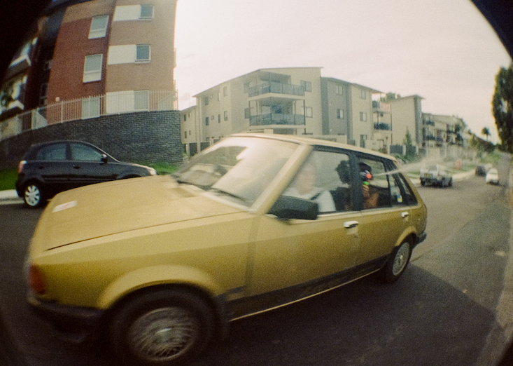 Fish-eye style photo of a yellow car driving past. The nozzle of a water gun can be seen spraying from the rear passenger window